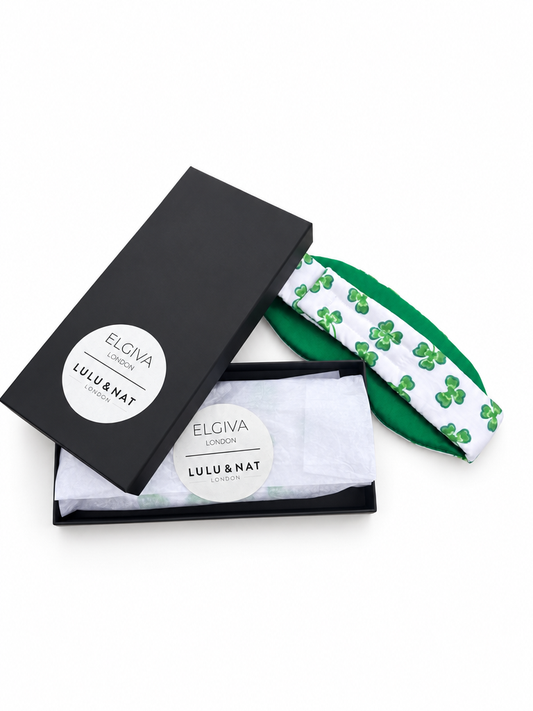 A Lulu & Nat ELGIVA London collaborative blackout sleep mask featuring green and white fabric outer made with Lulu and Nat's Lucky Clover cotton, an emerald green heavy silk lining, pure cotton inner wrapped in white tissue paper inside a black branded presentation box