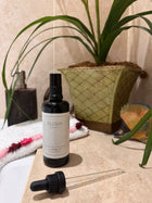A bottle of ELGIVA London Lavender Oil next to a bath with a black dropper pipette resting in front of it on a beige stone surface, with a potted plant and bathroom decor in the background
