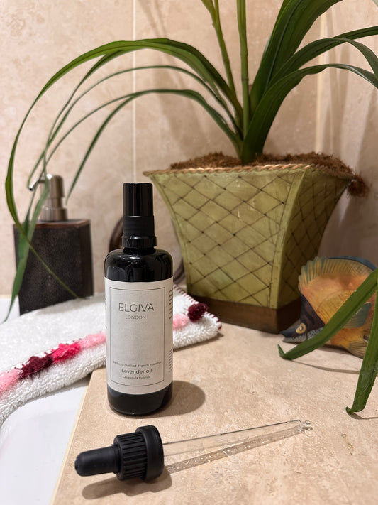 A bottle of ELGIVA London Lavender Oil next to a bath with a black dropper pipette resting in front of it on a beige stone surface, with a potted plant and bathroom decor in the background